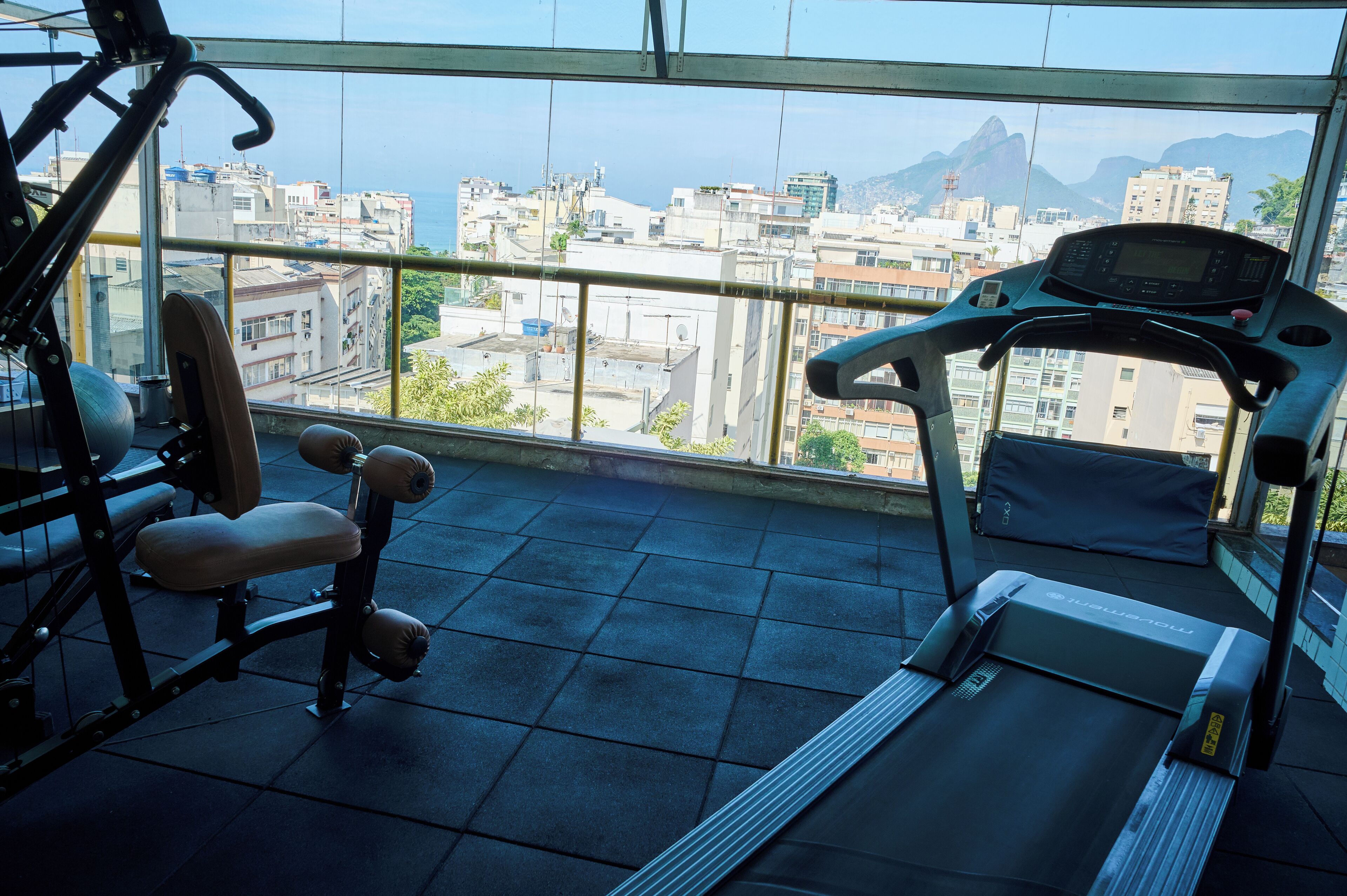 A gym with exercise equipment and a treadmill, overlooking a cityscape with buildings and mountains.