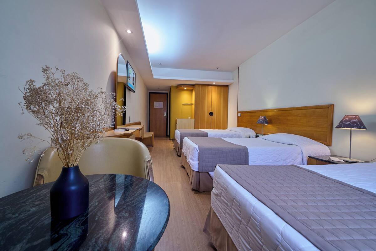 A hotel room with three beds, a desk with a vase of dried flowers, a wooden headboard, and a lamp.