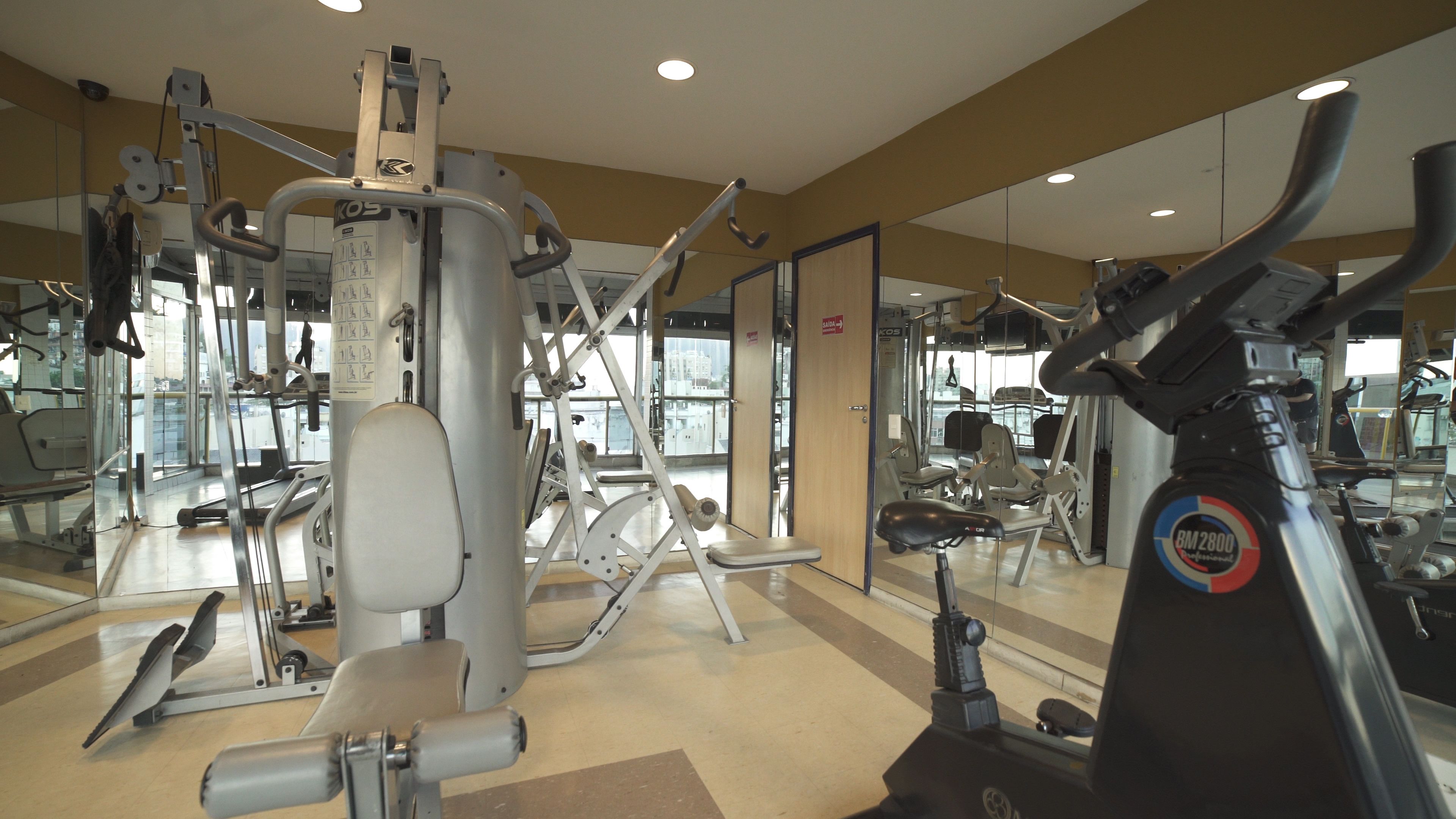 Une salle de sport équipée de divers appareils d’exercice, de miroirs et d’une porte donnant sur une autre pièce.