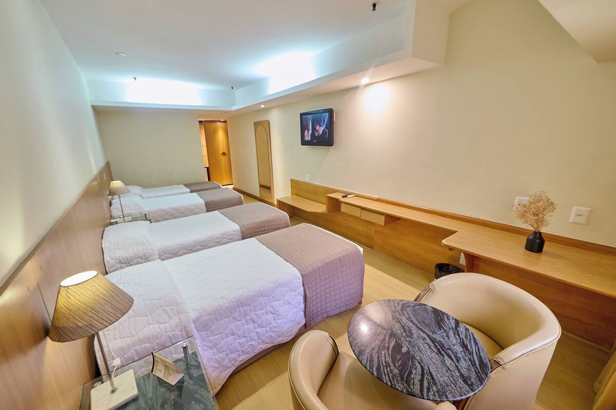 A hotel room with three beds, a desk, a chair, a TV, and a vase with dried flowers.