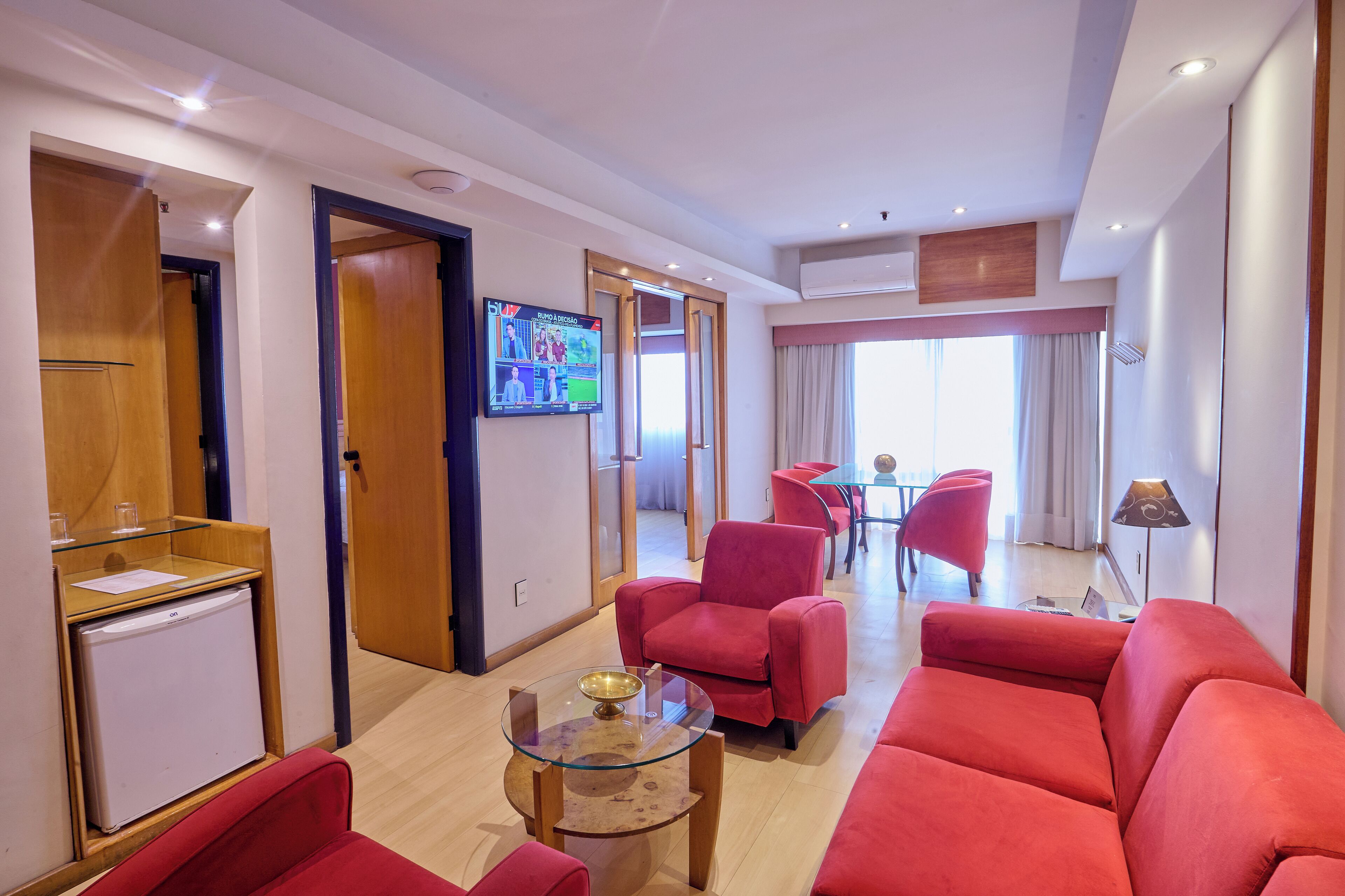 Une chambre d’hôtel moderne avec un canapé rouge, une table basse en verre, un téléviseur fixé au mur et un petit réfrigérateur.