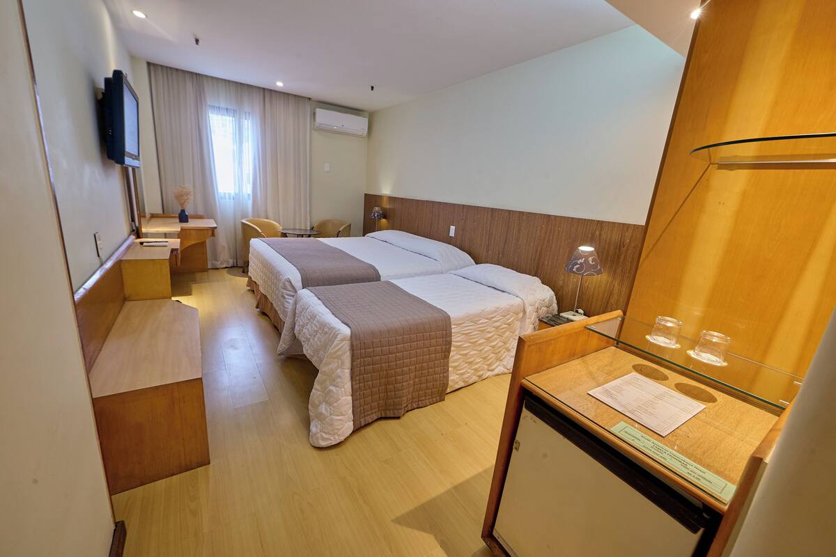 A hotel room with a bed, bedside tables, a desk, a chair, a TV, and a window with curtains.