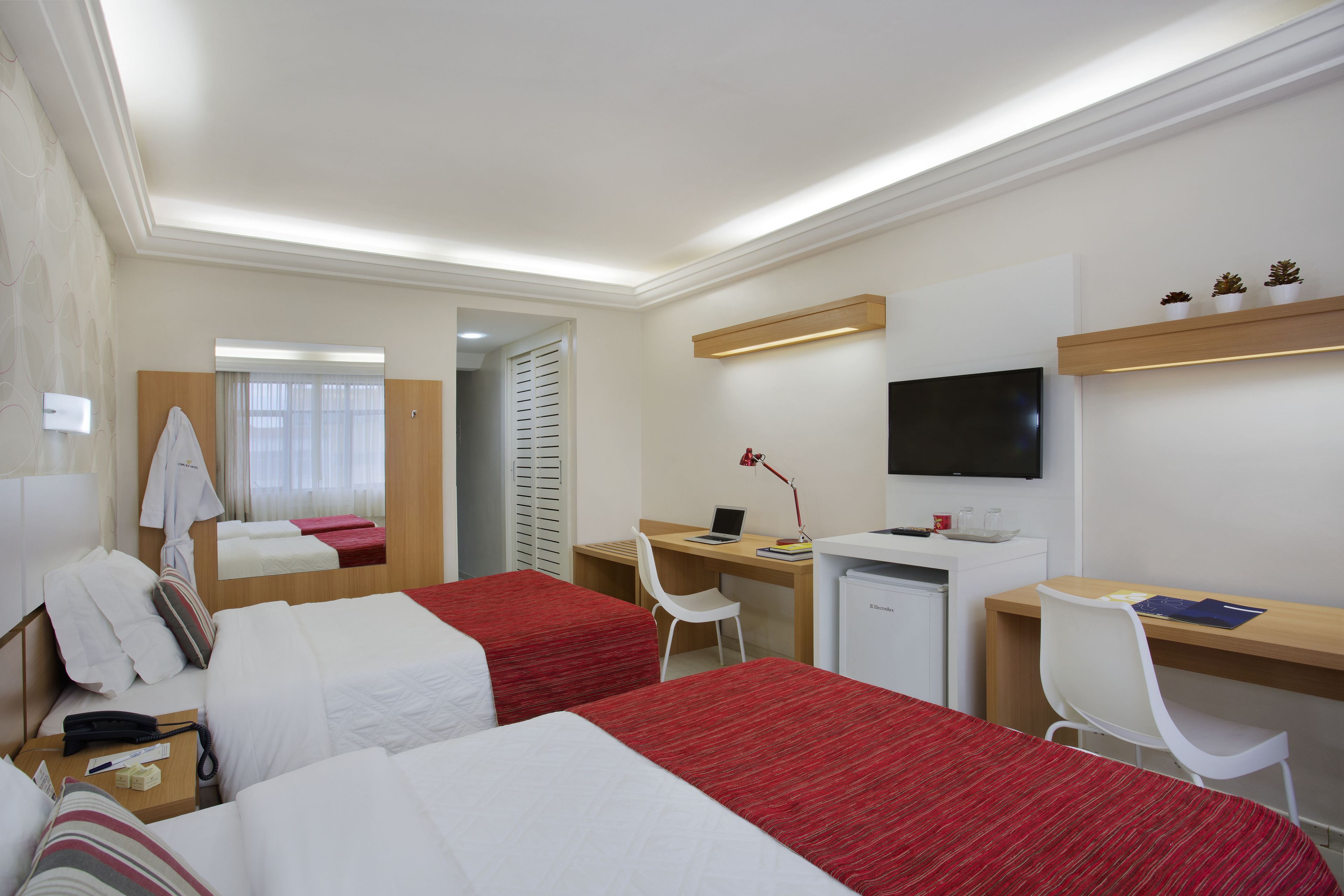 Quarto de hotel com duas camas, uma escrivaninha, uma cadeira, uma televisão e um espelho.