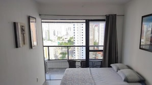 A bedroom with a bed, a chair, a window with a view of buildings, and a curtain.