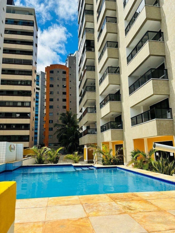 A swimming pool surrounded by high-rise buildings with balconies, a clear sky, and some palm trees.