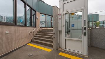 Entrada de un edificio con una escalera, una puerta con señal de accesibilidad para sillas de ruedas y un letrero azul de estacionamiento para discapacitados.
