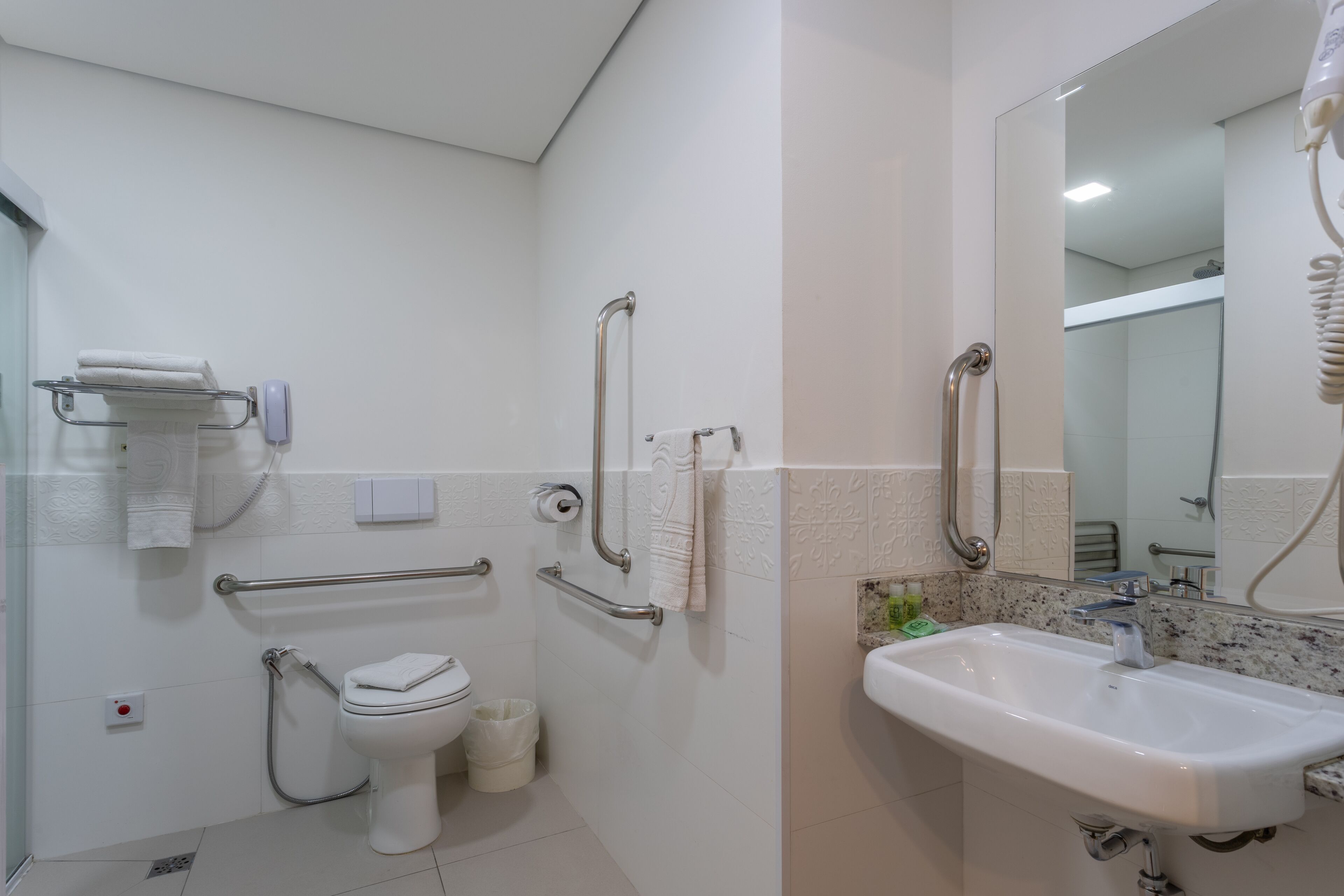 A modern bathroom with a toilet, bidet, and sink. It includes grab bars, a towel rack, and a mirror.