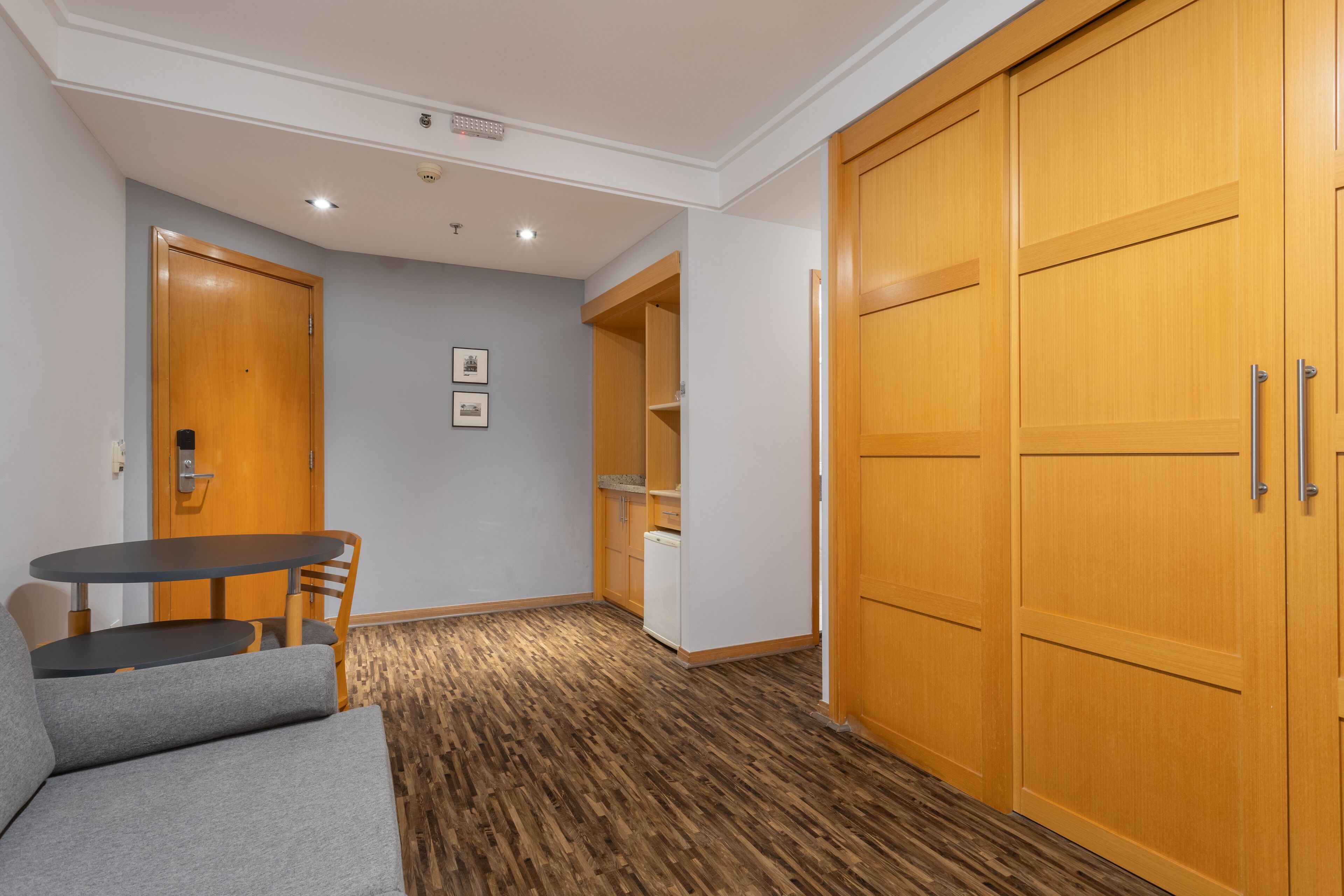 Una habitación con un armario de madera, un sofá gris, una mesita pequeña y una pared blanca con dos cuadros enmarcados.