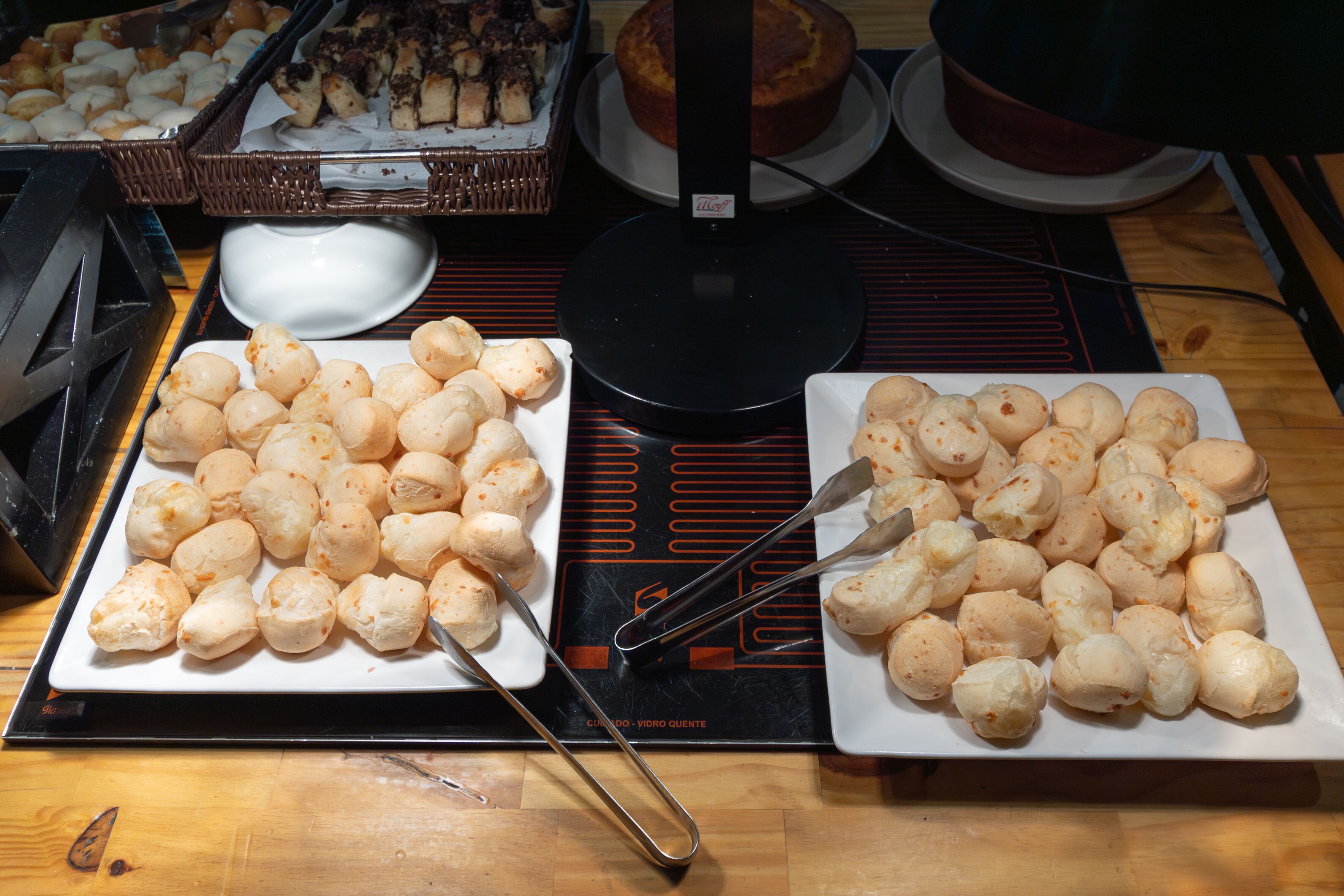 Uma mesa de buffet com diversos alimentos, incluindo uma travessa de pães e uma travessa de bolinhos de queijo.