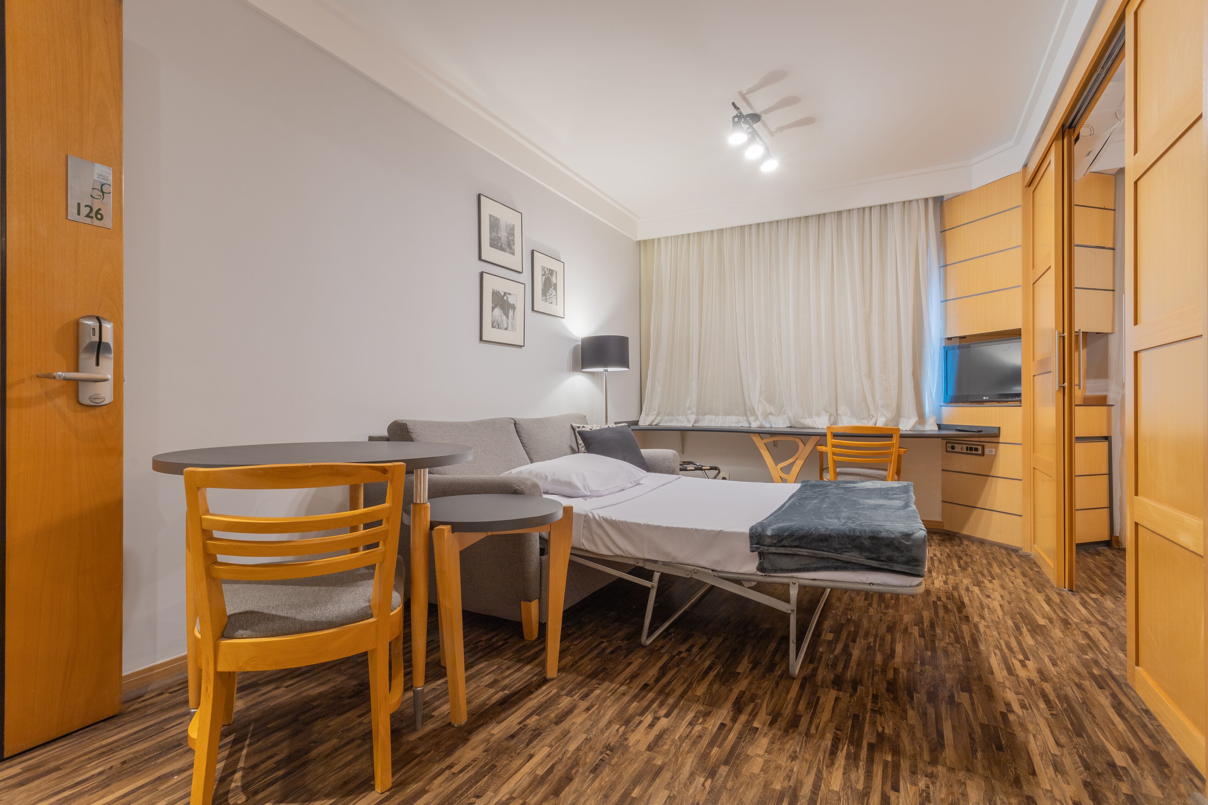 A compact room with a bed, a small table, a chair, a desk, and a wardrobe.