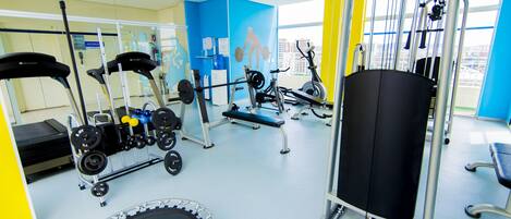 A gym with exercise equipment, a trampoline, and a view of buildings through large windows.
