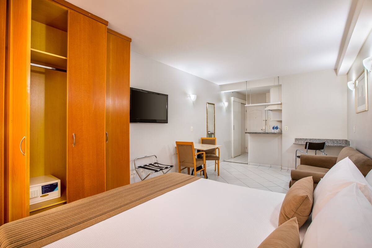A hotel room with a bed, a desk, a chair, a wardrobe, a television, and a kitchenette.
