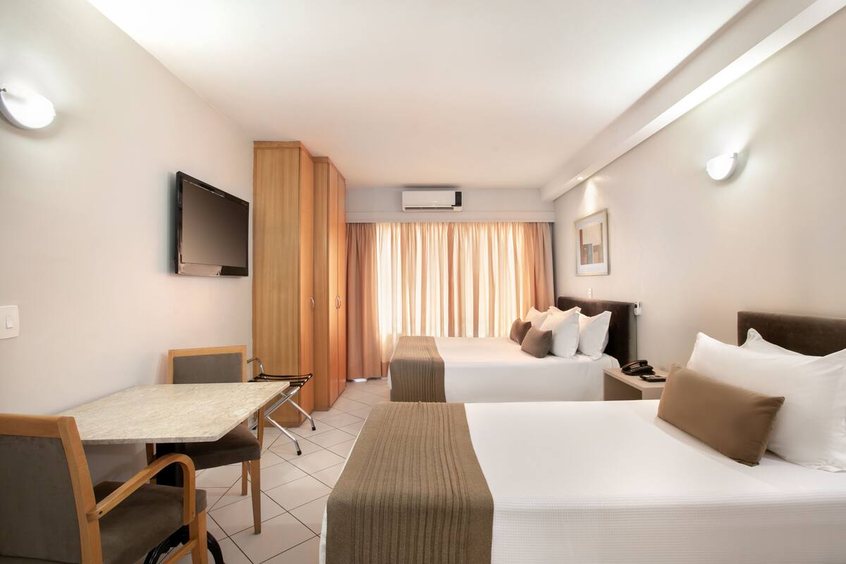 A hotel room with two beds, a TV, a desk, and a window with curtains.