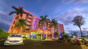 A multi-story hotel with a modern design, featuring a prominent entrance and a parking area with several cars and motorcycles. - Falls Galli Hotel (Foz do Iguaçu)