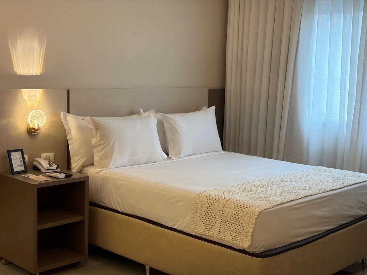 Quarto de hotel com uma cama, uma mesinha de cabeceira com telefone e luminária, e uma janela com cortinas.