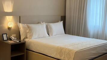 Quarto de hotel com uma cama, uma mesinha de cabeceira com telefone e luminária, e uma janela com cortinas.