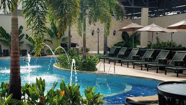 Ein Poolbereich im Innenbereich mit Liegestühlen und Sonnenschirmen, umgeben von Pflanzen und einer gefliesten Poolanlage.