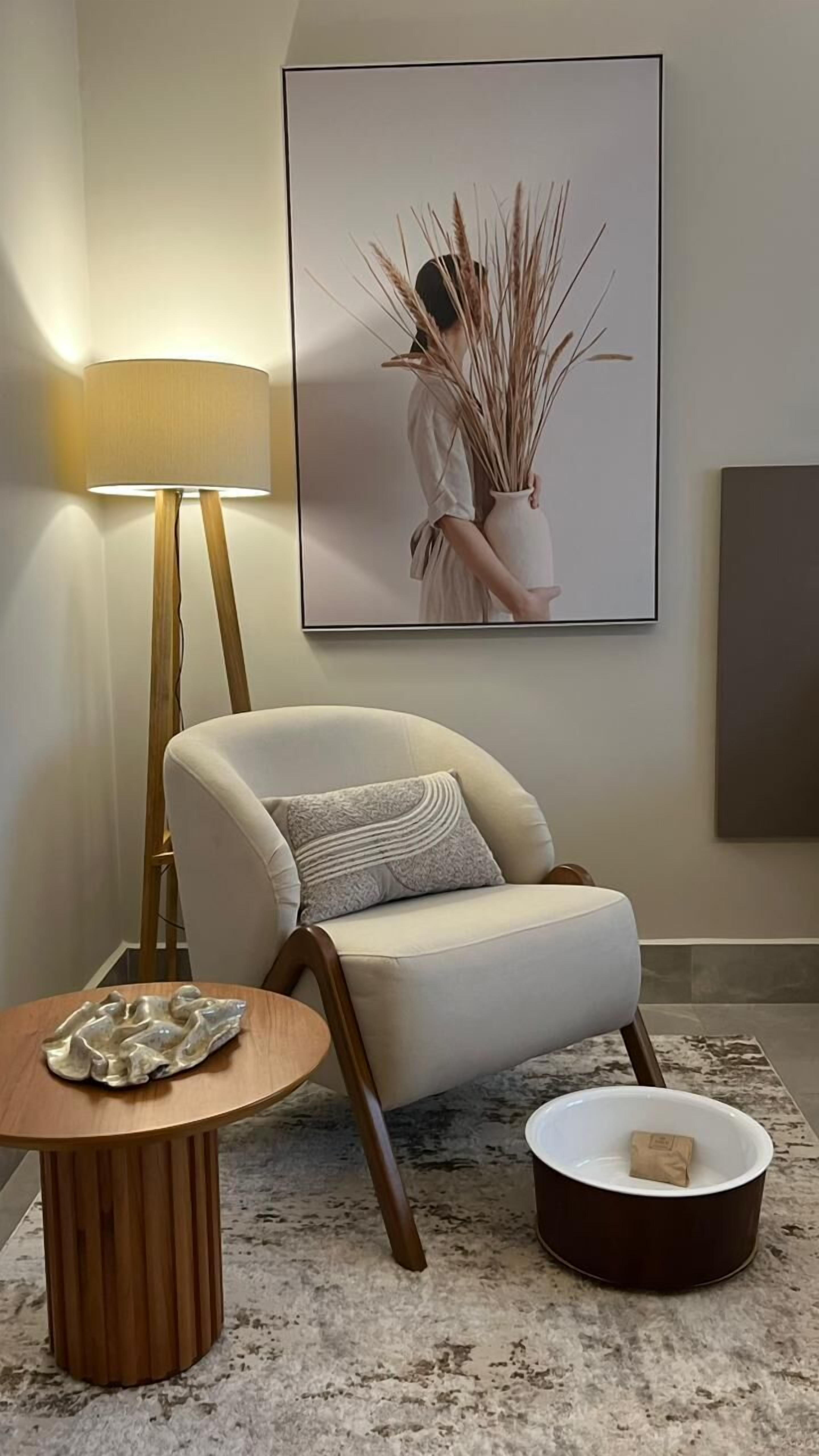 A modern living room with a white armchair, a round wooden side table, a floor lamp, and a framed artwork on the wall.