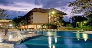 Un hotel con piscina, varios balcones y fachada iluminada al anochecer.