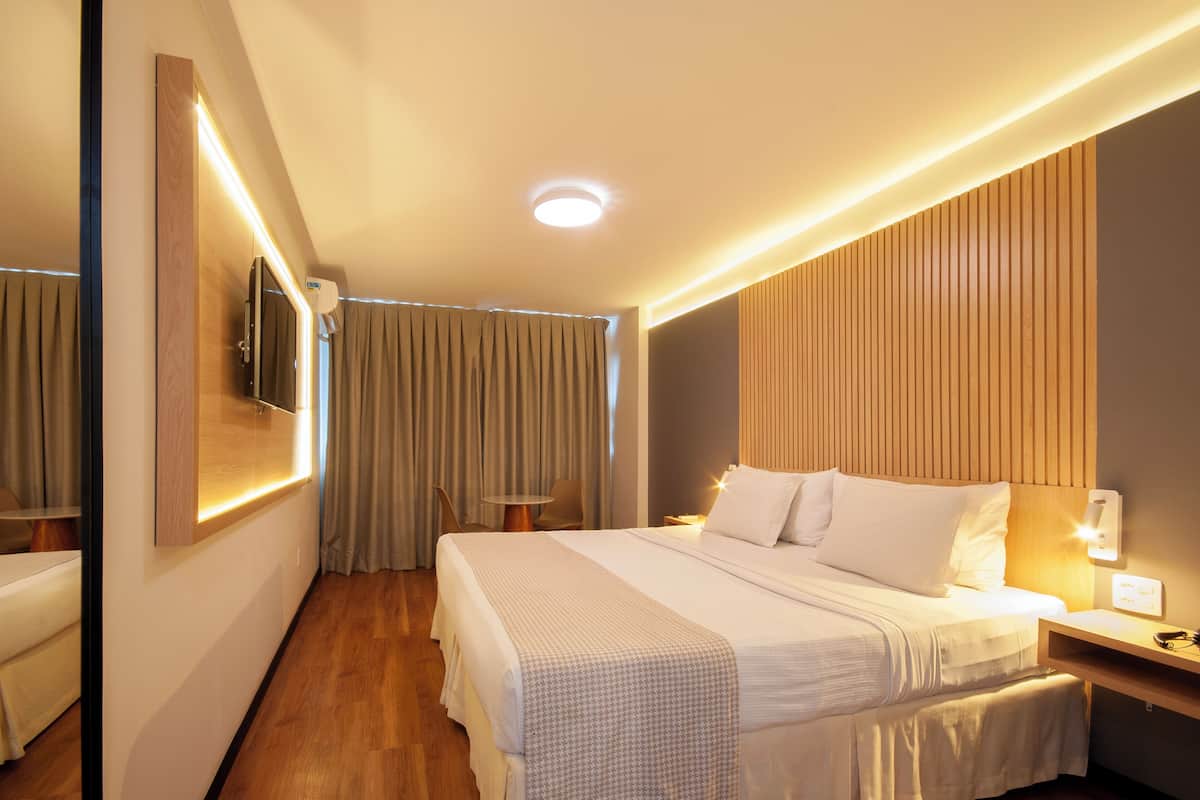 Een hotelkamer met een bed, nachtkastje, lamp, spiegel en een klein tafeltje met een stoel.