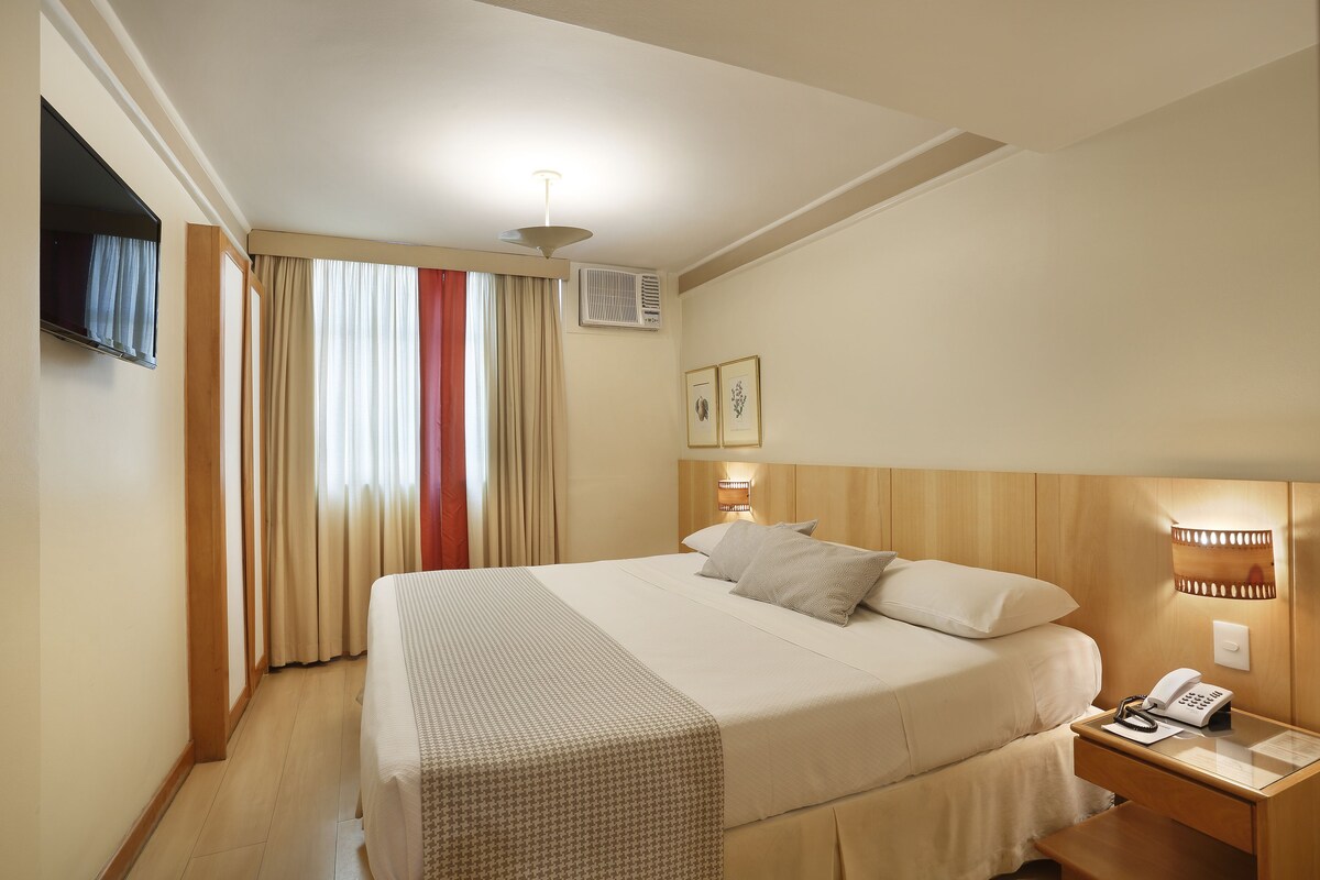 Een hotelkamer met een bed, nachtkastje, lamp en een aan de muur bevestigde televisie.
