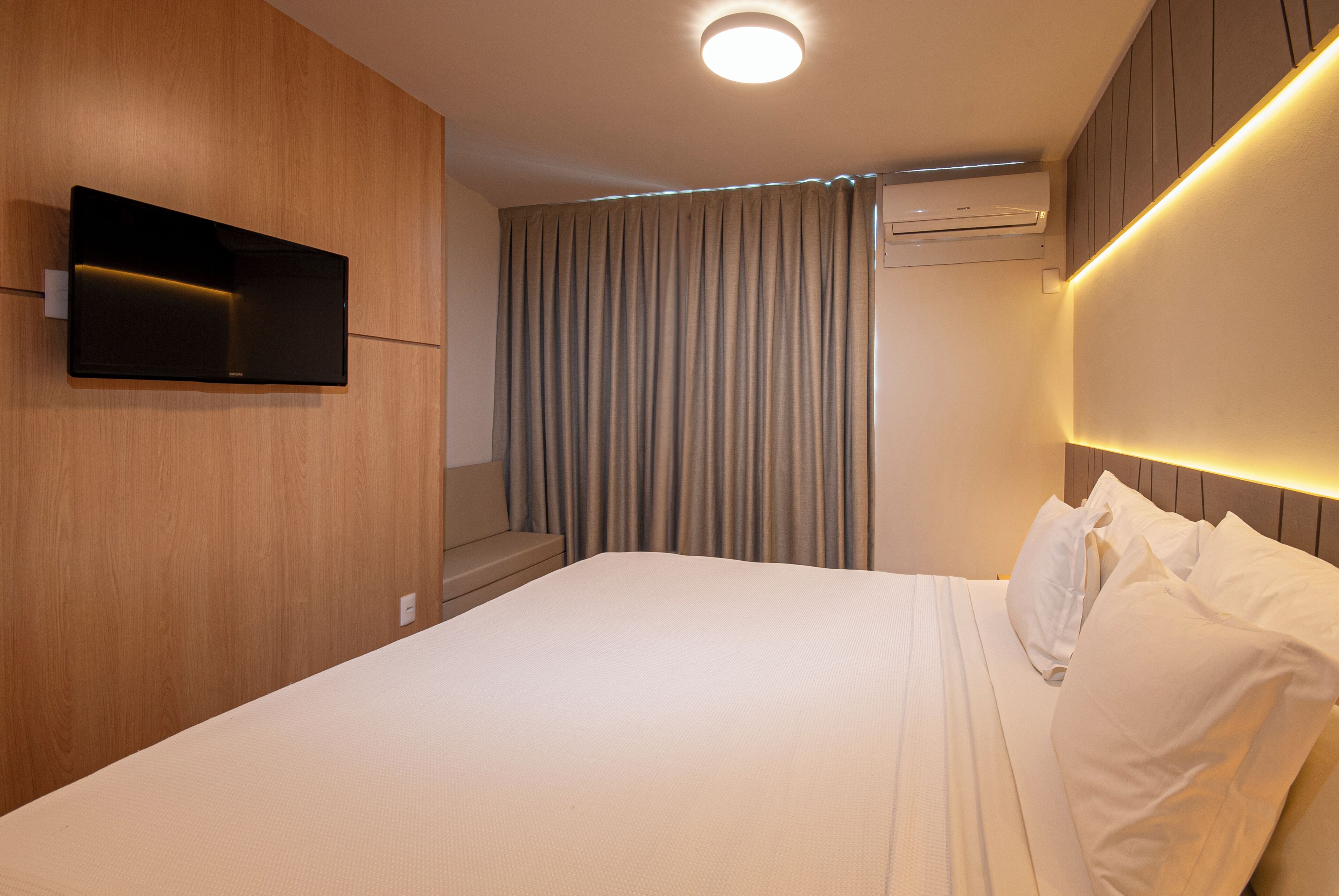 A hotel room with a bed, a mounted TV, an air conditioning unit, and a curtain.