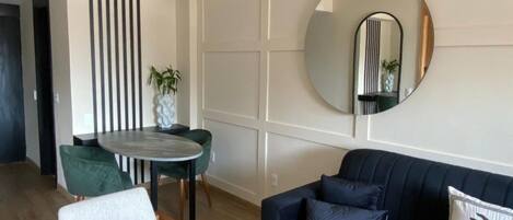 A modern living room with a dark blue sofa, a round mirror, and a dining area with a small table and chairs.