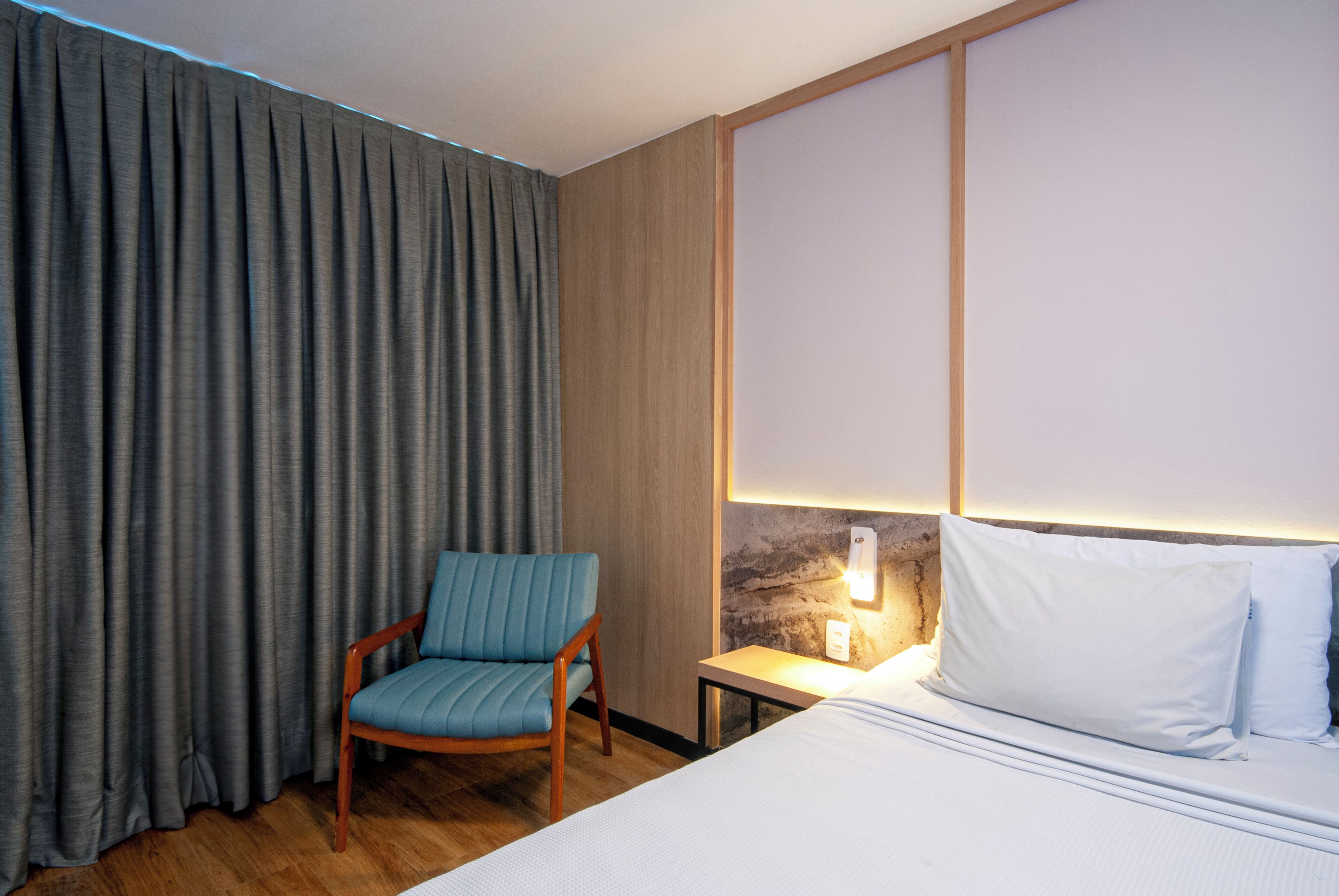 Quarto de hotel com uma cama, uma cadeira, um criado-mudo e uma cortina.