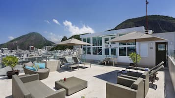 Terraço no telhado com mobiliário de vime, guarda-sóis e vista para a paisagem urbana e montanhas.