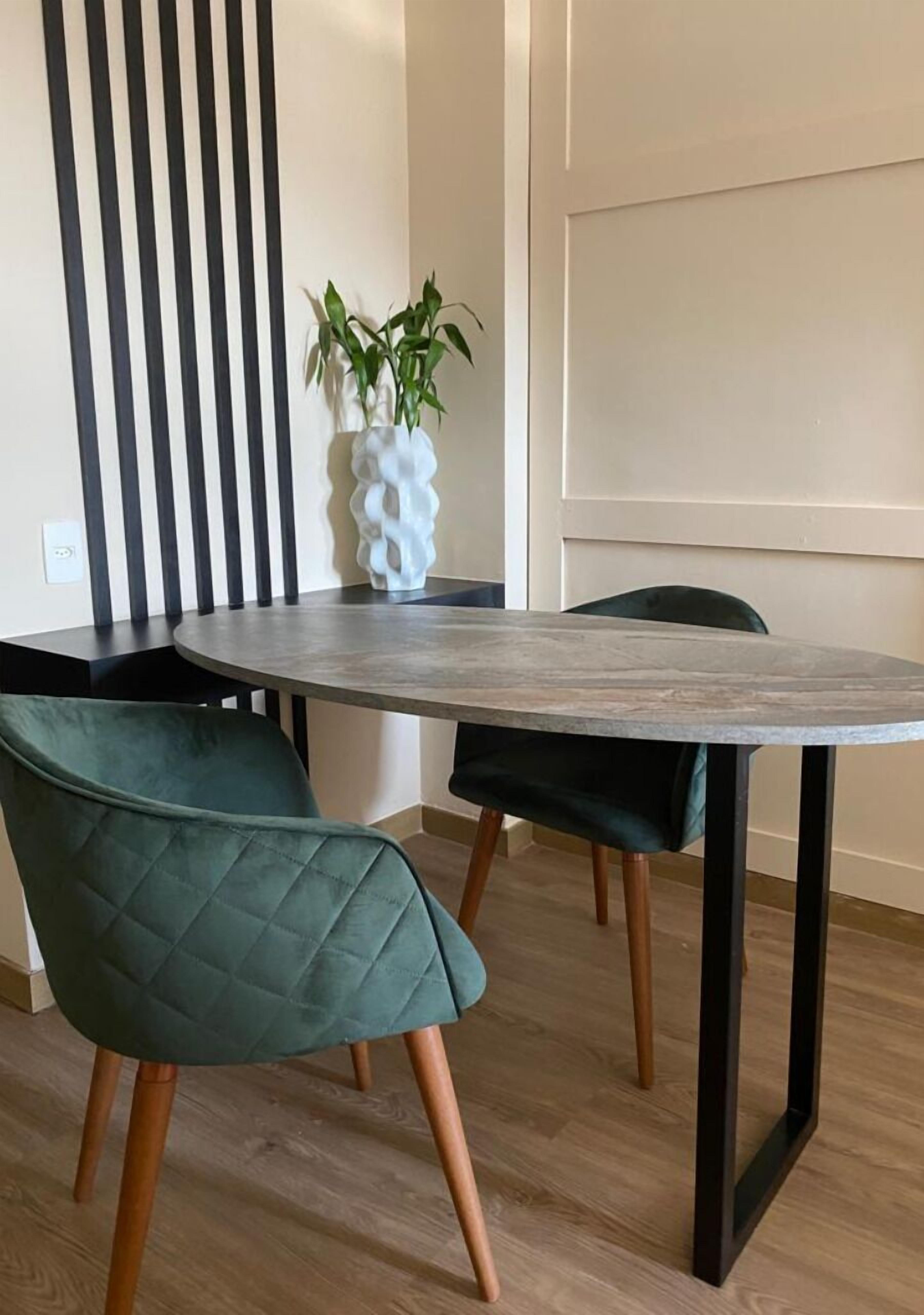 A dining area with a round table, two green upholstered chairs, a vase with a plant, and a decorative wall panel.