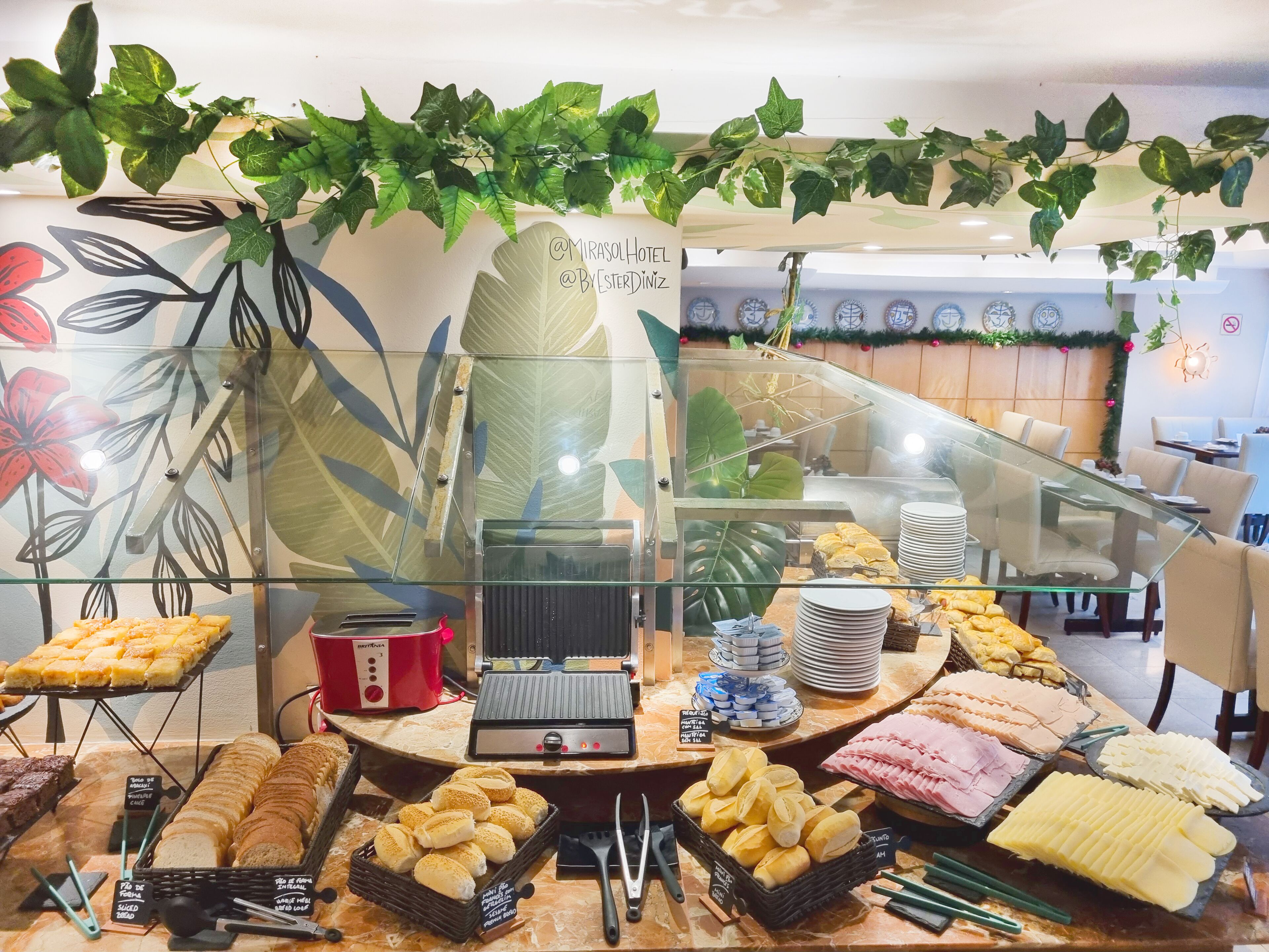 A buffet setup with various food items, a coffee machine, and a decorative plant overhead.