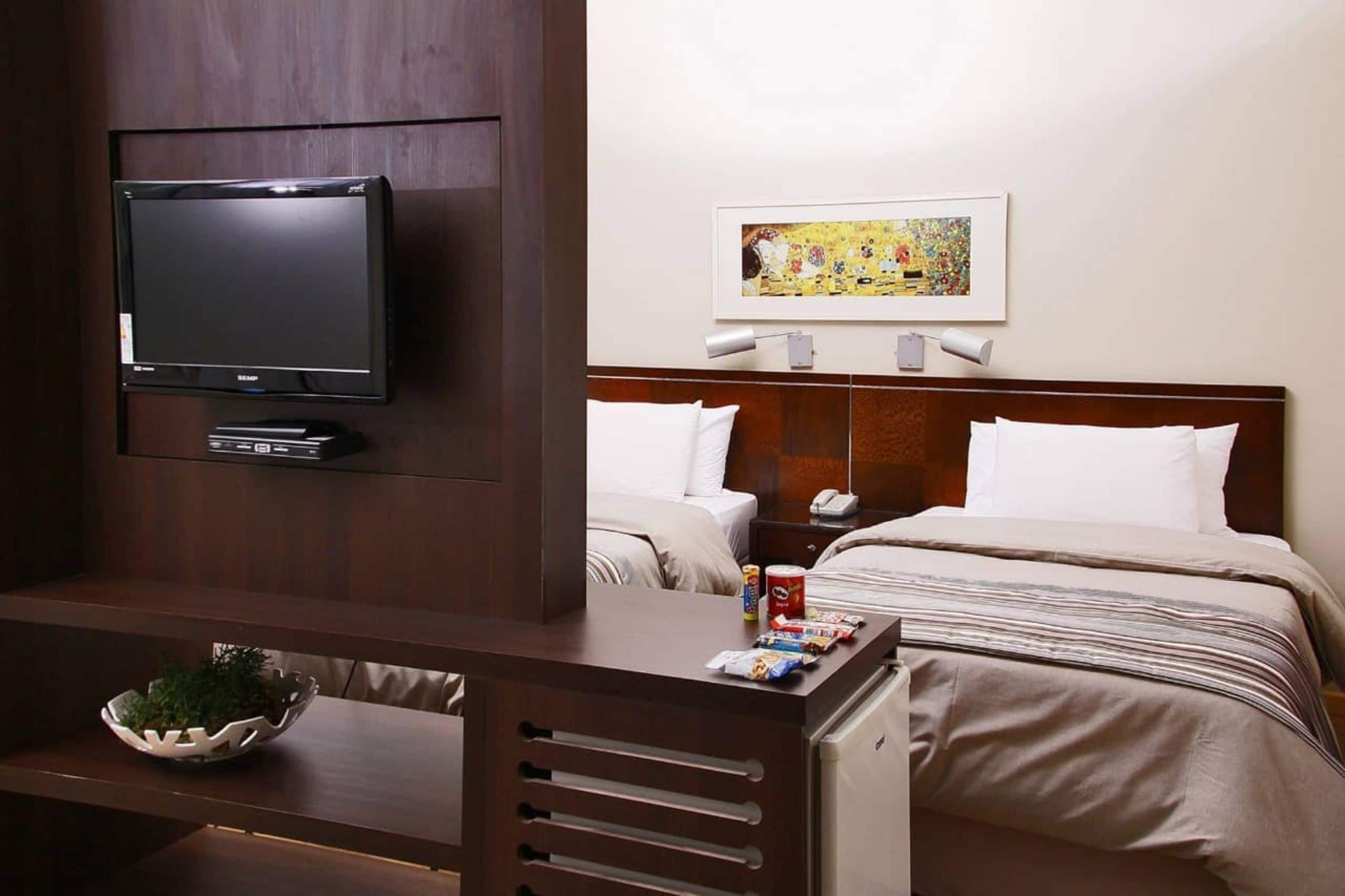 A hotel room with a bed, a television, a painting, and a small table with snacks.