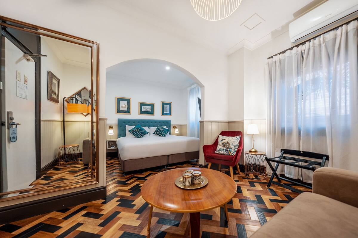 Ein Hotelzimmer mit Bett, einem roten Sessel, einem Holztisch und einem Spiegel, mit Blick auf den Flur.