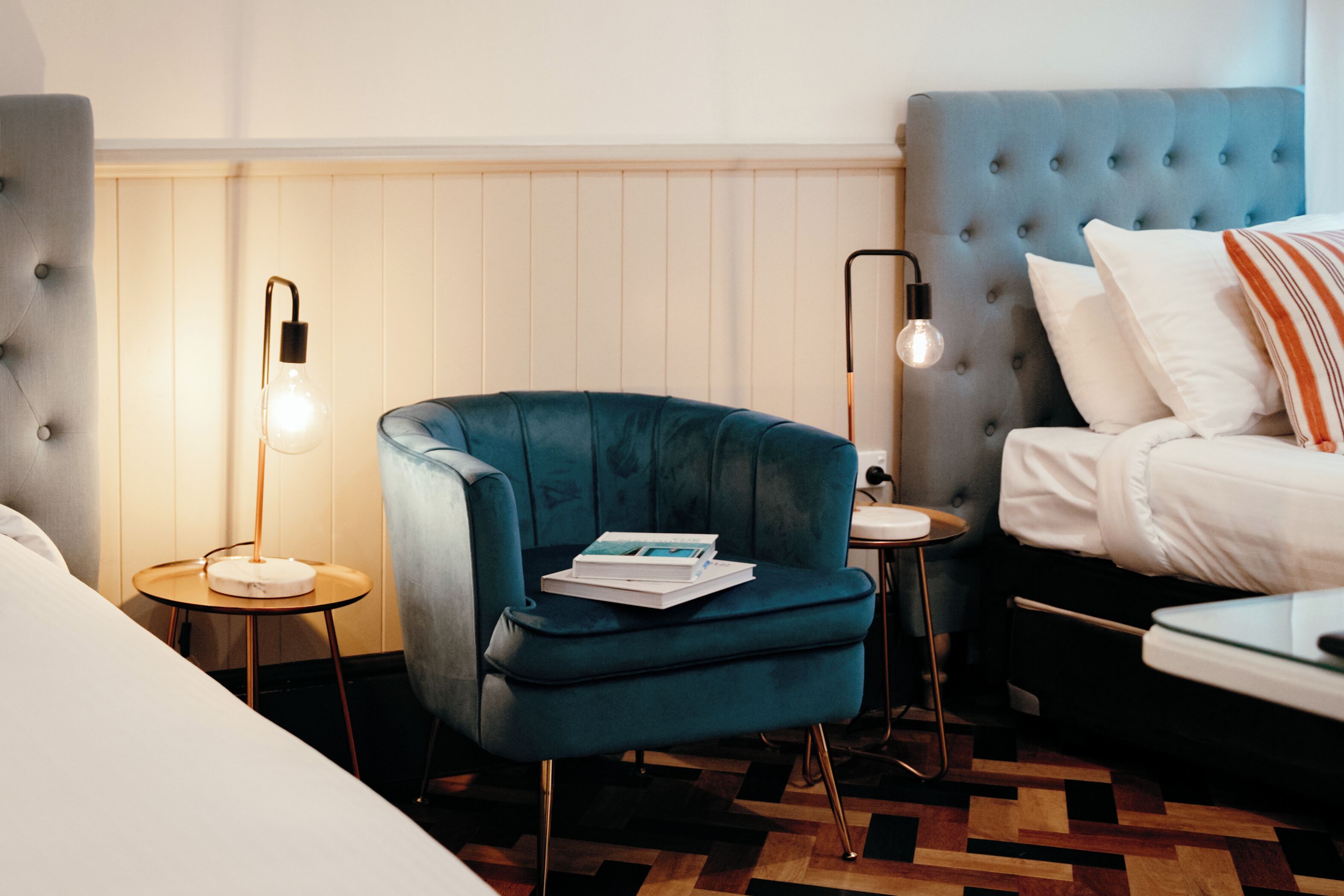 Ein modernes Hotelzimmer mit einer gesteppten Kopfteil, einem türkisfarbenen Sessel, einem kleinen runden Tisch mit einer Lampe und einem Bett mit weißer Bettwäsche.