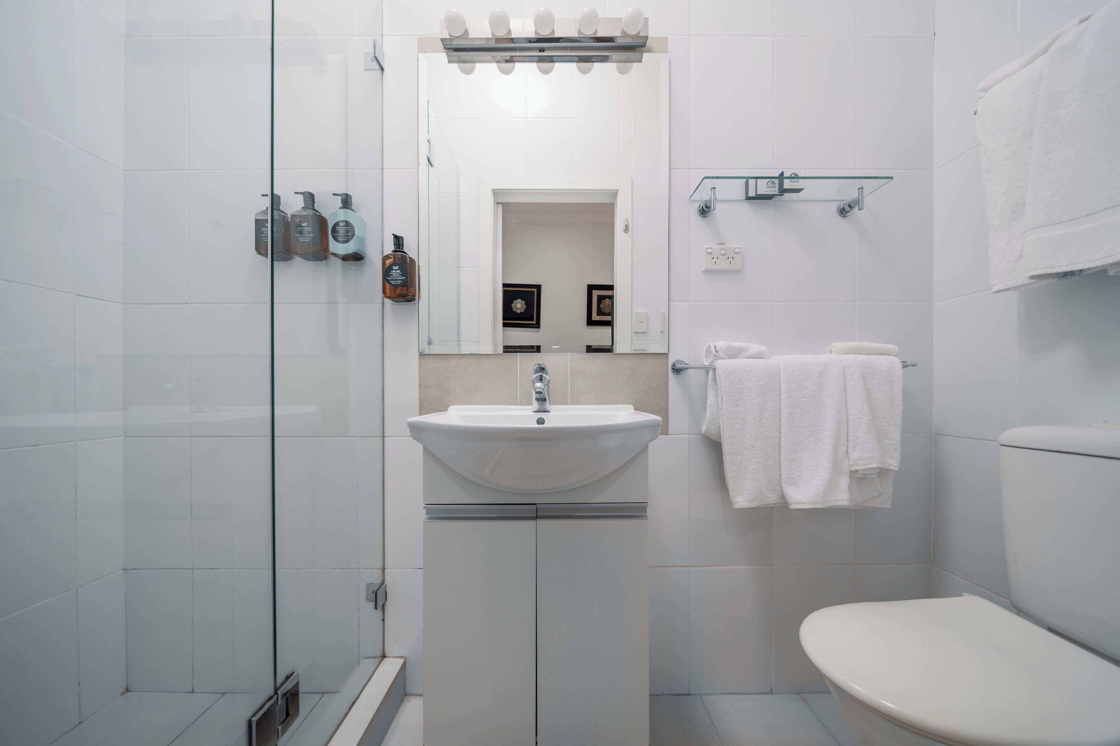 A modern bathroom with a white sink, mirror, and toilet. There are toiletries on the wall and a glass shower enclosure.