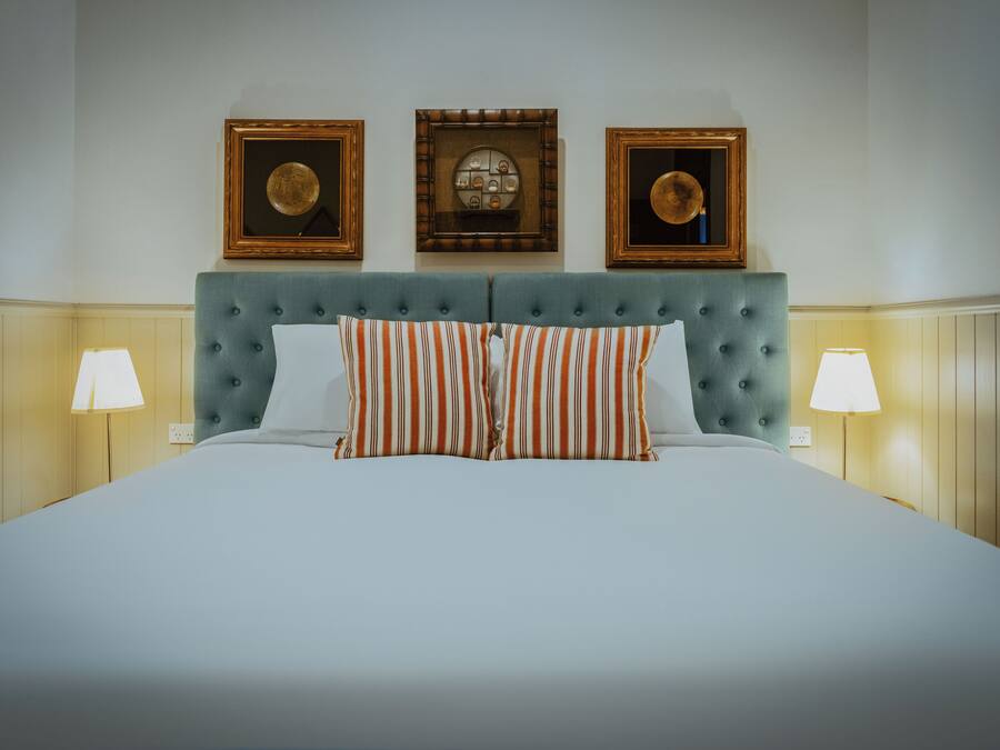 Una cama impecablemente arreglada con almohadillas a rayas, un cabecero capitonado y dos lámparas de pared a cada lado.