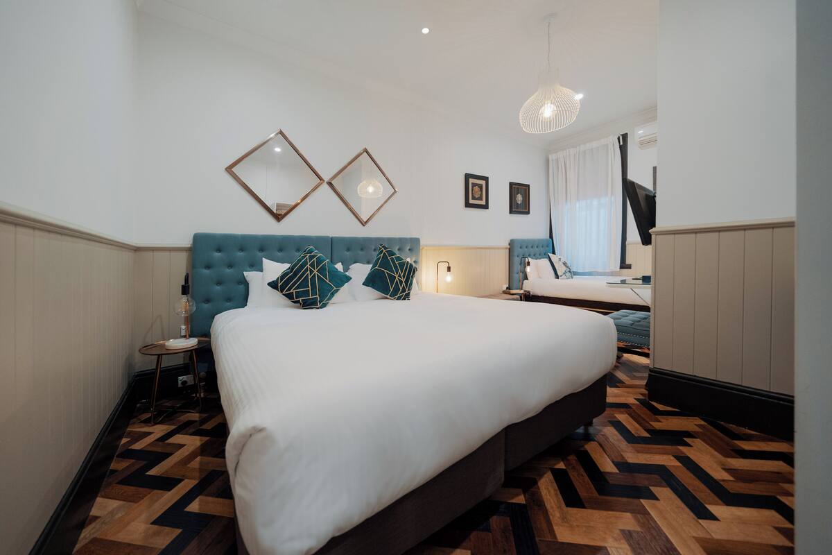 Ein Hotelzimmer mit einem großen Bett, einem blauen Kopfteil, zwei Wandspiegeln, einem Holzboden im Fischgrätmuster und einer hängenden Leuchte.