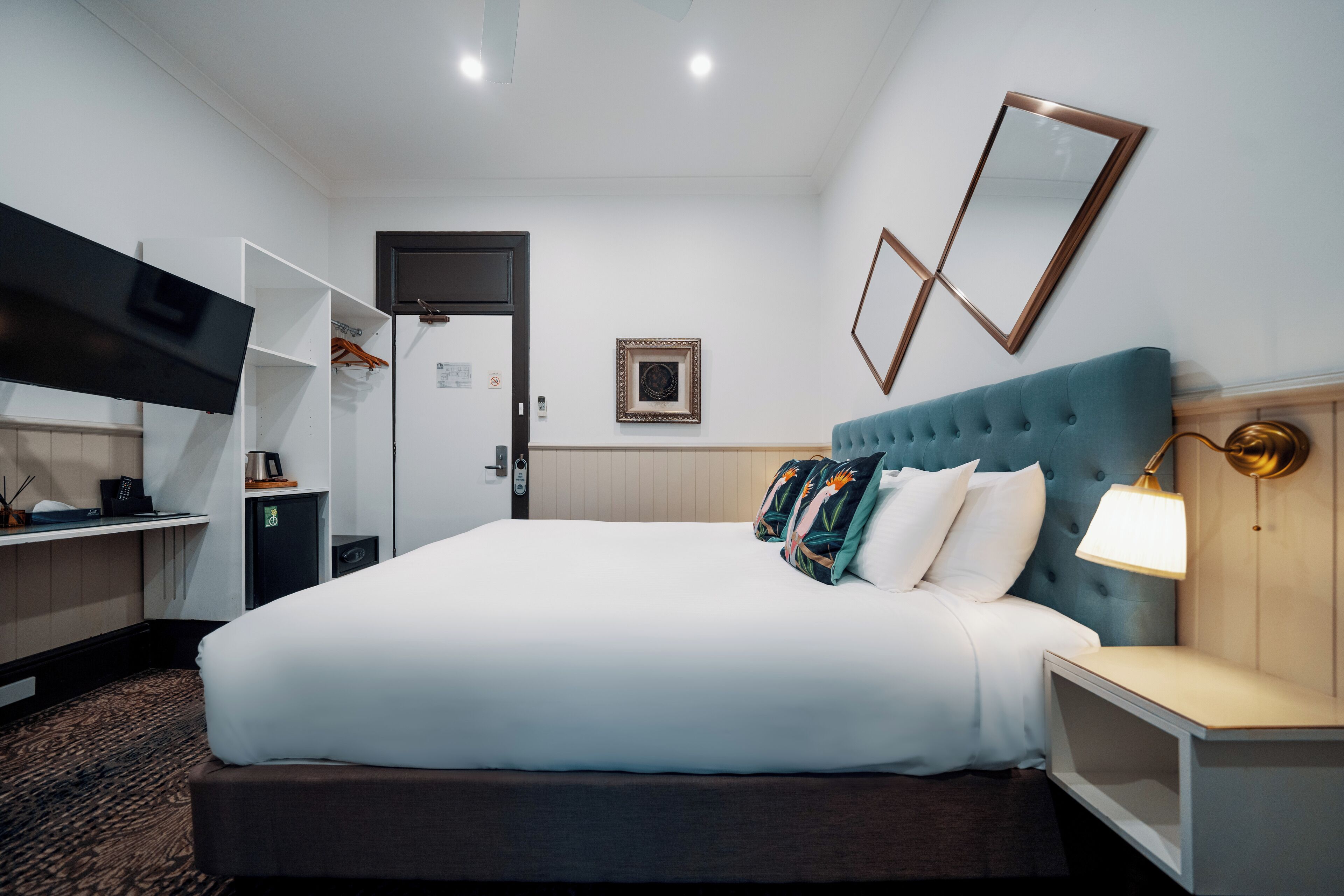 A hotel room with a large bed, a TV mounted on the wall, a bedside table with a lamp, and a wardrobe.