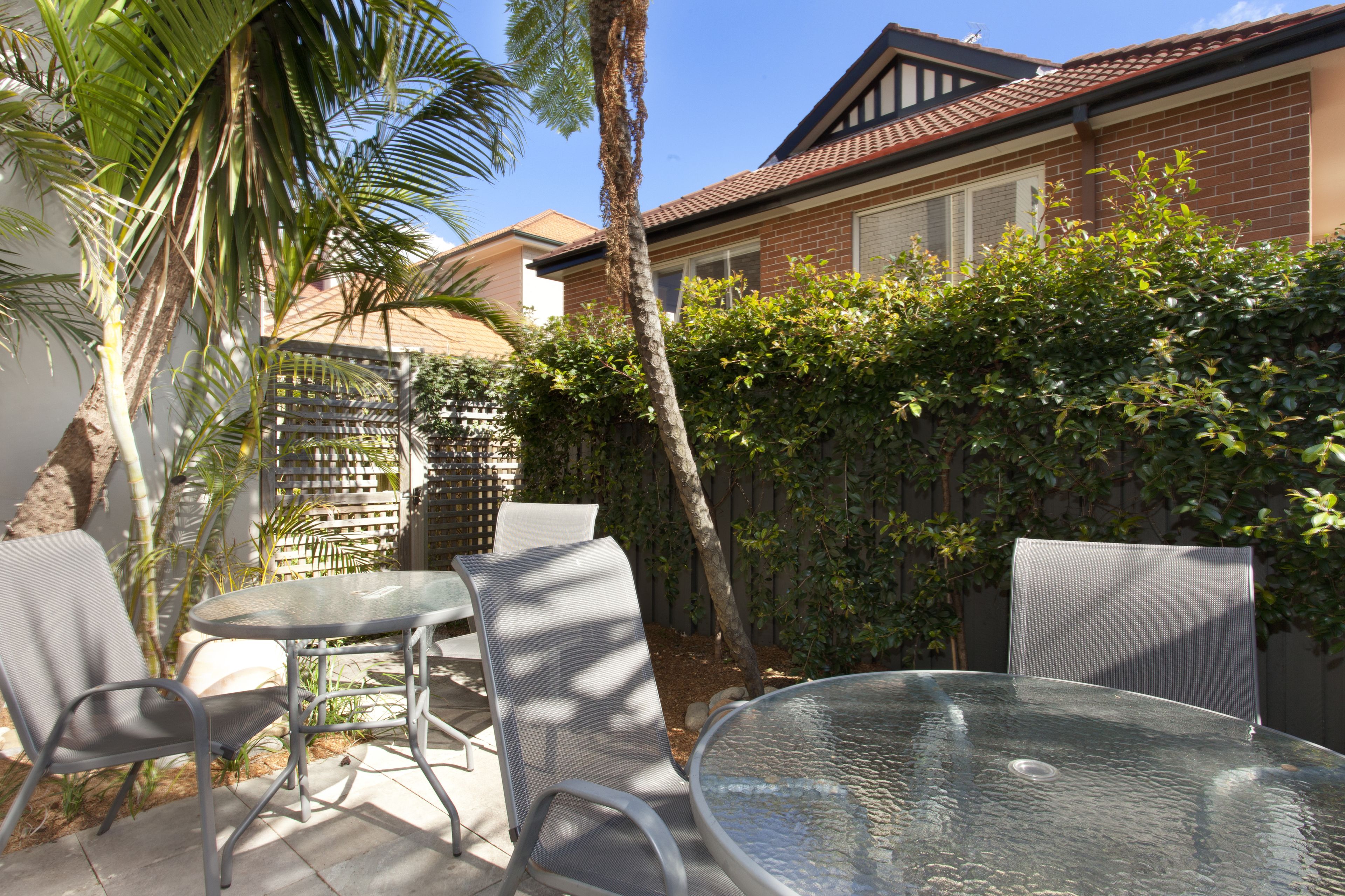 A patio with a glass table and metal chairs, surrounded by a brick house and a wooden fence.