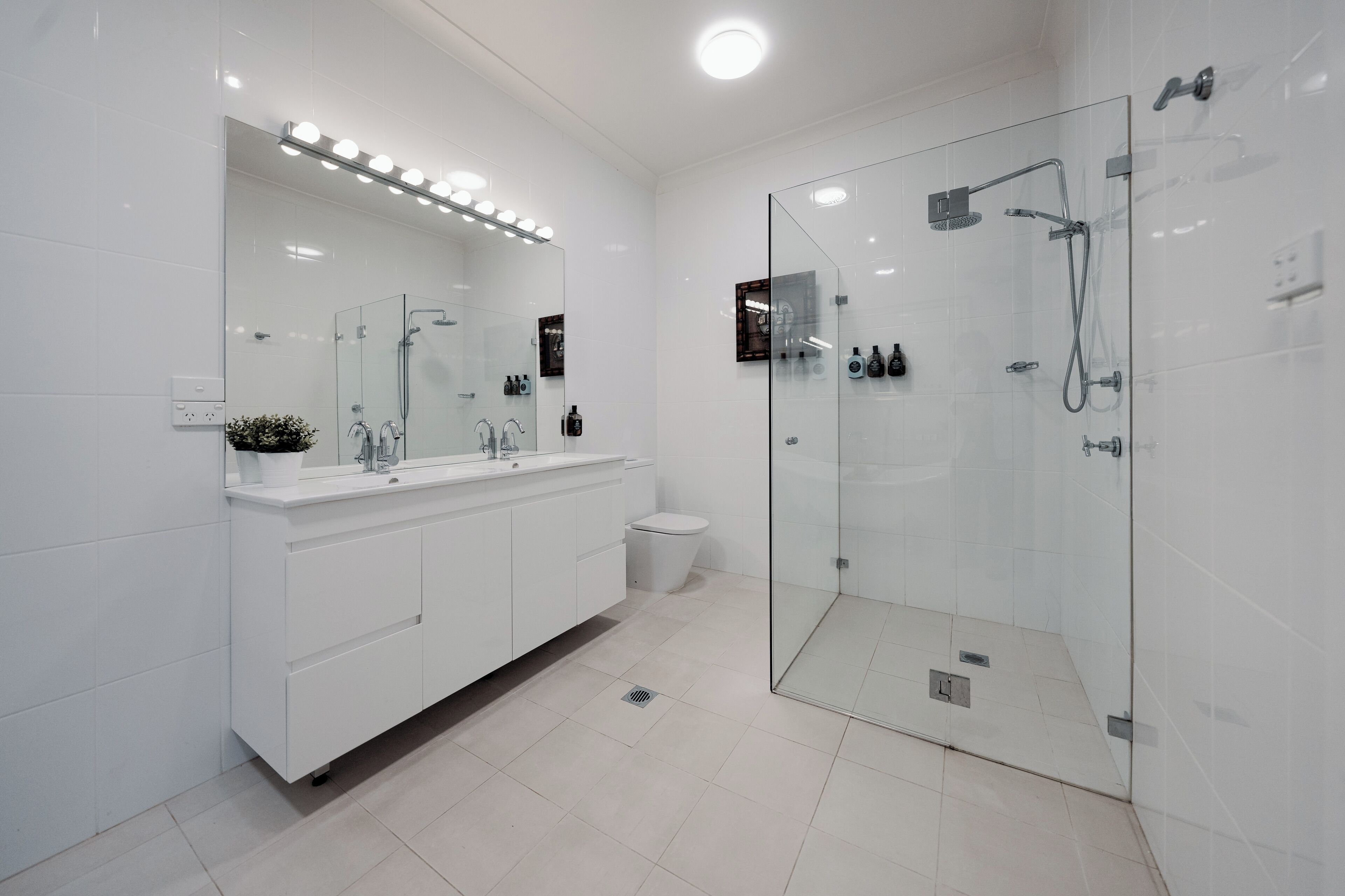 A modern bathroom with a glass-enclosed shower, white cabinetry, and a large mirror with lights above.
