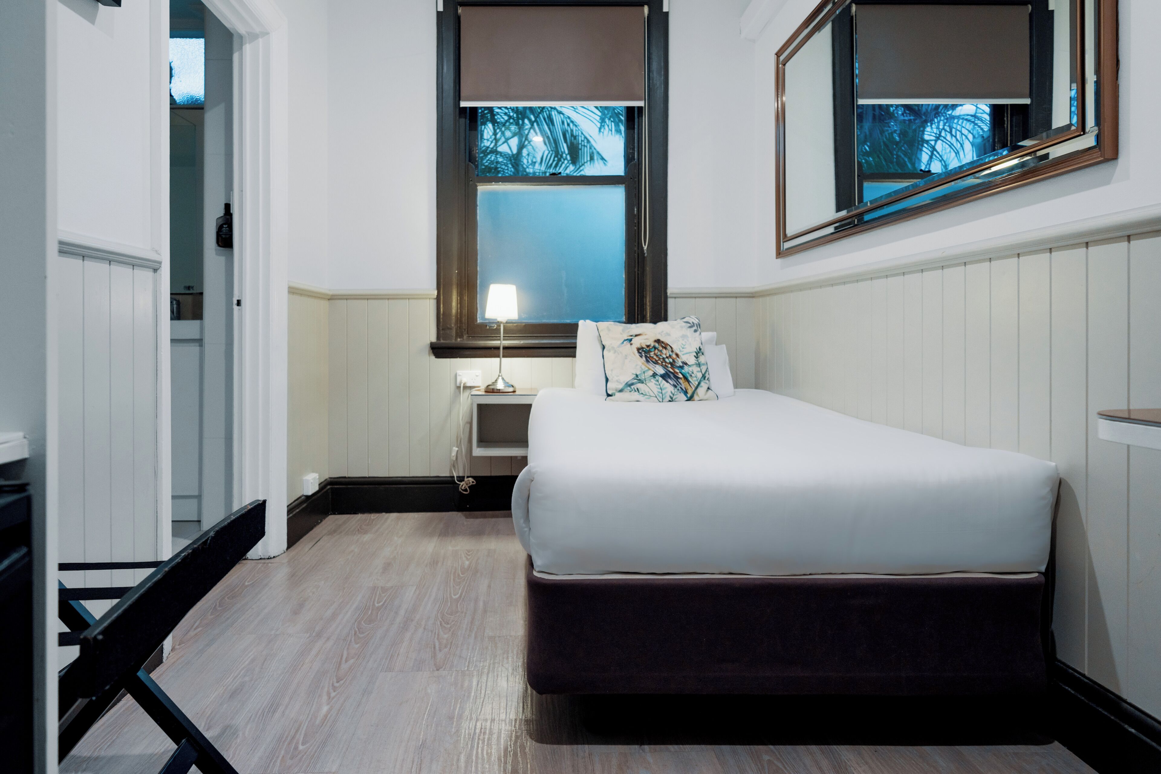 A bedroom with a bed, bedside table, lamp, and a window with a view of palm trees.