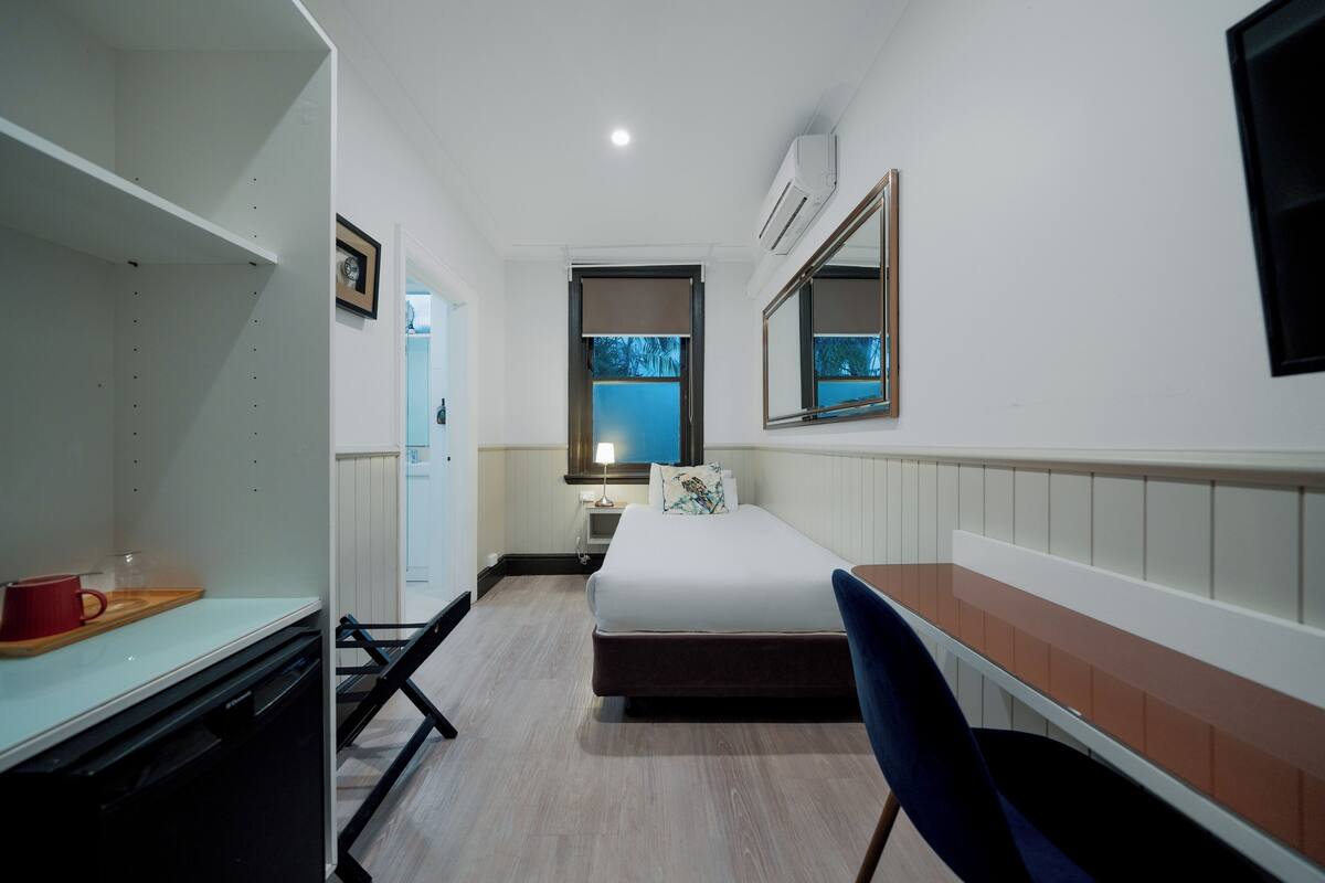 Ein Hotelzimmer mit Bett, Schreibtisch, Stuhl, einem kleinen Tisch mit roter Tasse, Lampe und Fenster mit Blick auf Bäume.