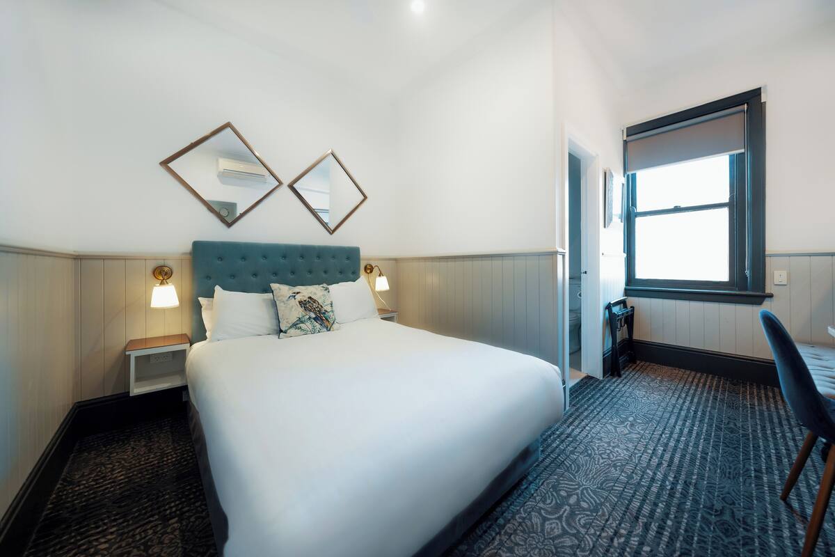 Ein Hotelzimmer mit einem großen Bett, zwei Nachttischen mit Lampen, einem Fenster mit Jalousien und einem Sessel.