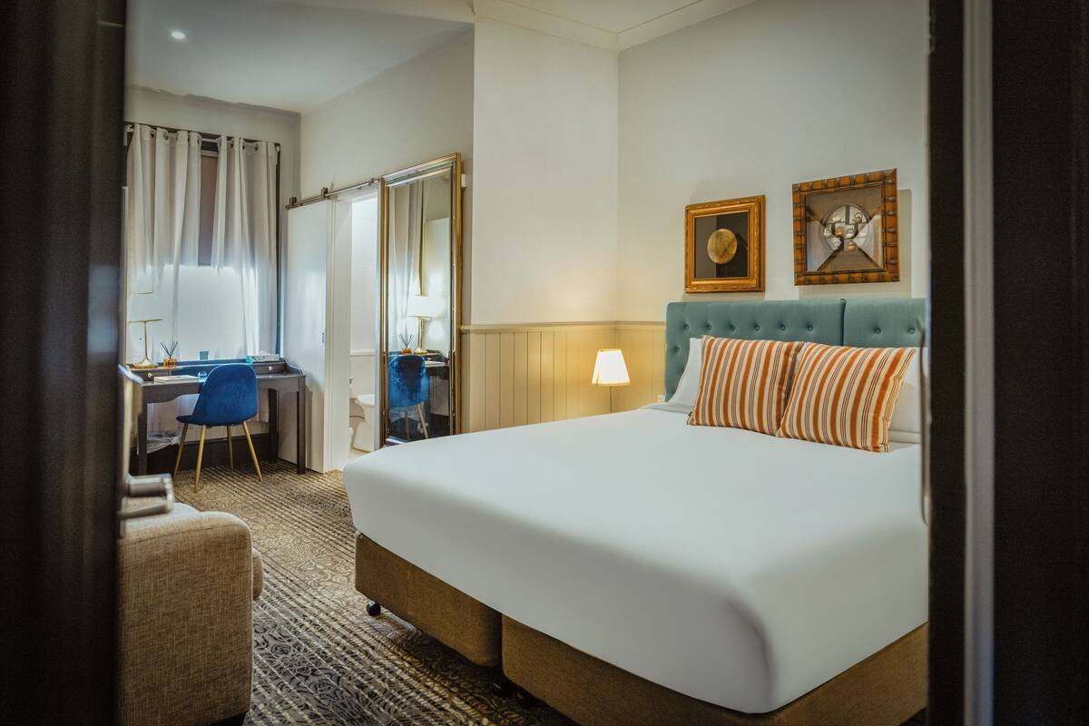 Ein Hotelzimmer mit einem großen Bett, einem Schreibtisch mit Stuhl, einem Spiegel und zwei gerahmten Bildern an der Wand.