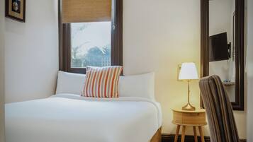 Habitación de hotel con una cama grande, una mesita de noche con lámpara, una silla, una ventana con vistas y un espejo en la pared.