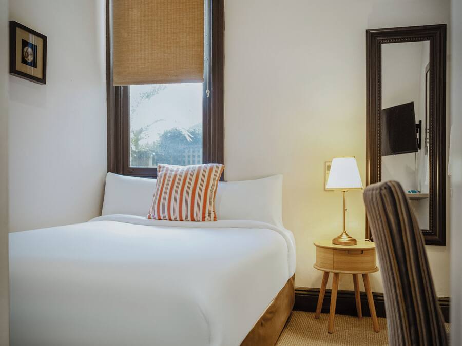 Habitación de hotel con una cama grande, una mesita de noche con lámpara, una silla, una ventana con vistas y un espejo en la pared.
