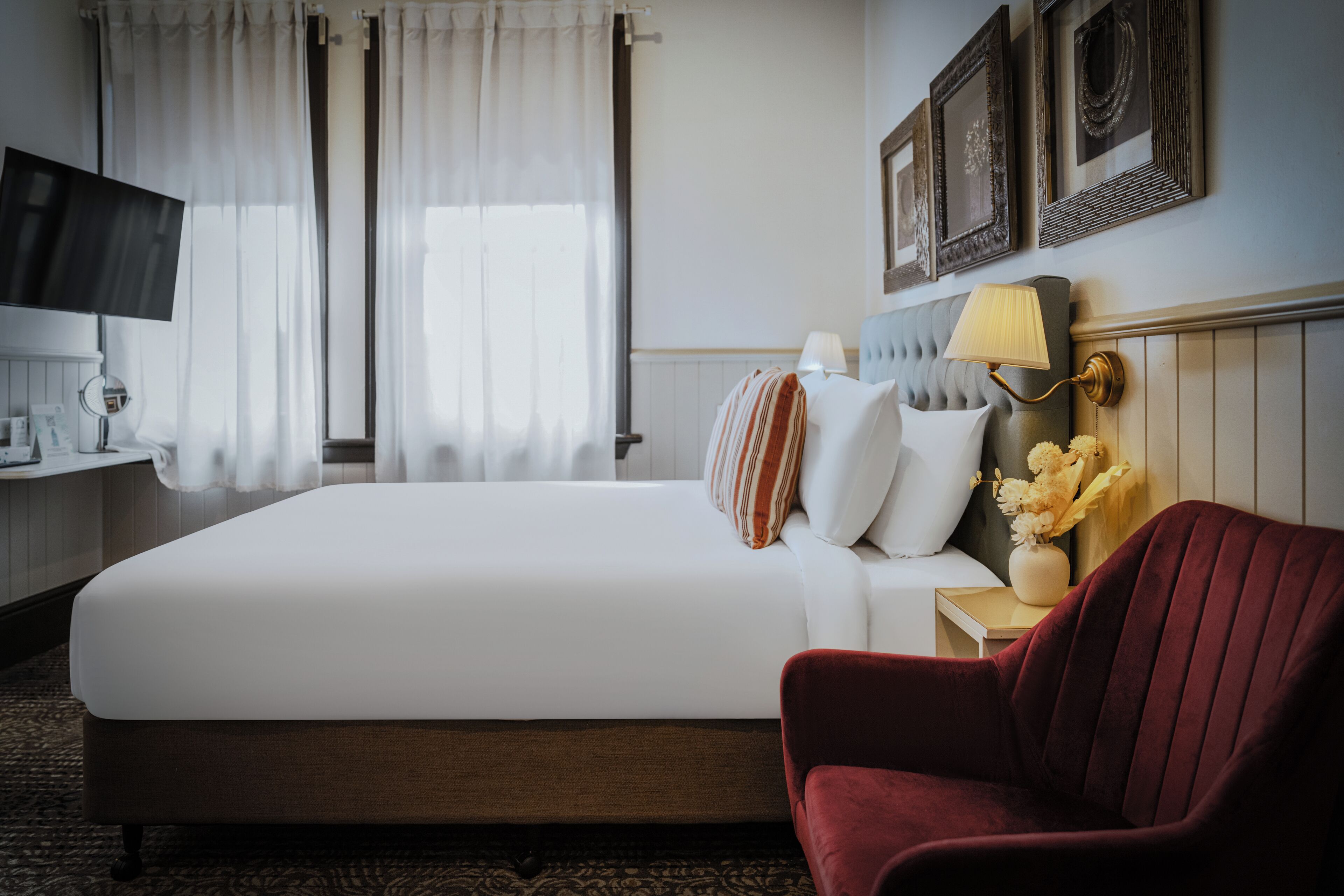 Ein Hotelzimmer mit einem großen Bett, einem roten Sessel, einem Nachttisch mit Lampe und einem an der Wand befestigten Fernseher.