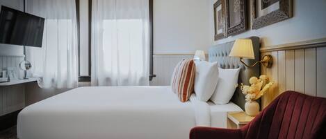 Habitación de hotel con una cama grande, un sillón rojo, una mesita de noche con lámpara y un televisor montado en la pared.