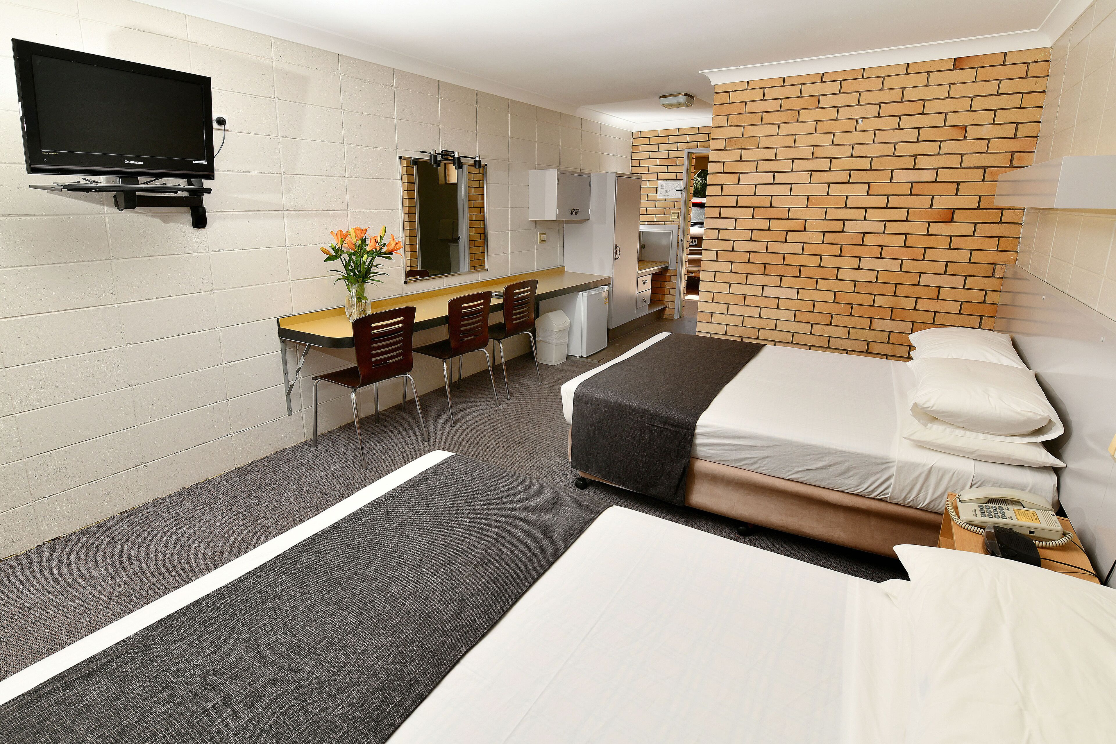 A hotel room with a bed, a desk with chairs, a television, and a brick-patterned wall.