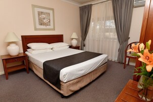 Una cama bien hecha con cabecera de madera, dos mesitas de noche con lámparas, un cuadro en la pared y un jarrón con flores sobre una mesita auxiliar.